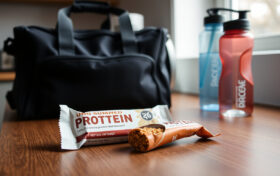 Kaksi merkitöntä proteiinipatukkaa puiseen pöytään aseteltuna gym-laukun ja vesipullon vieressä keittiössä.